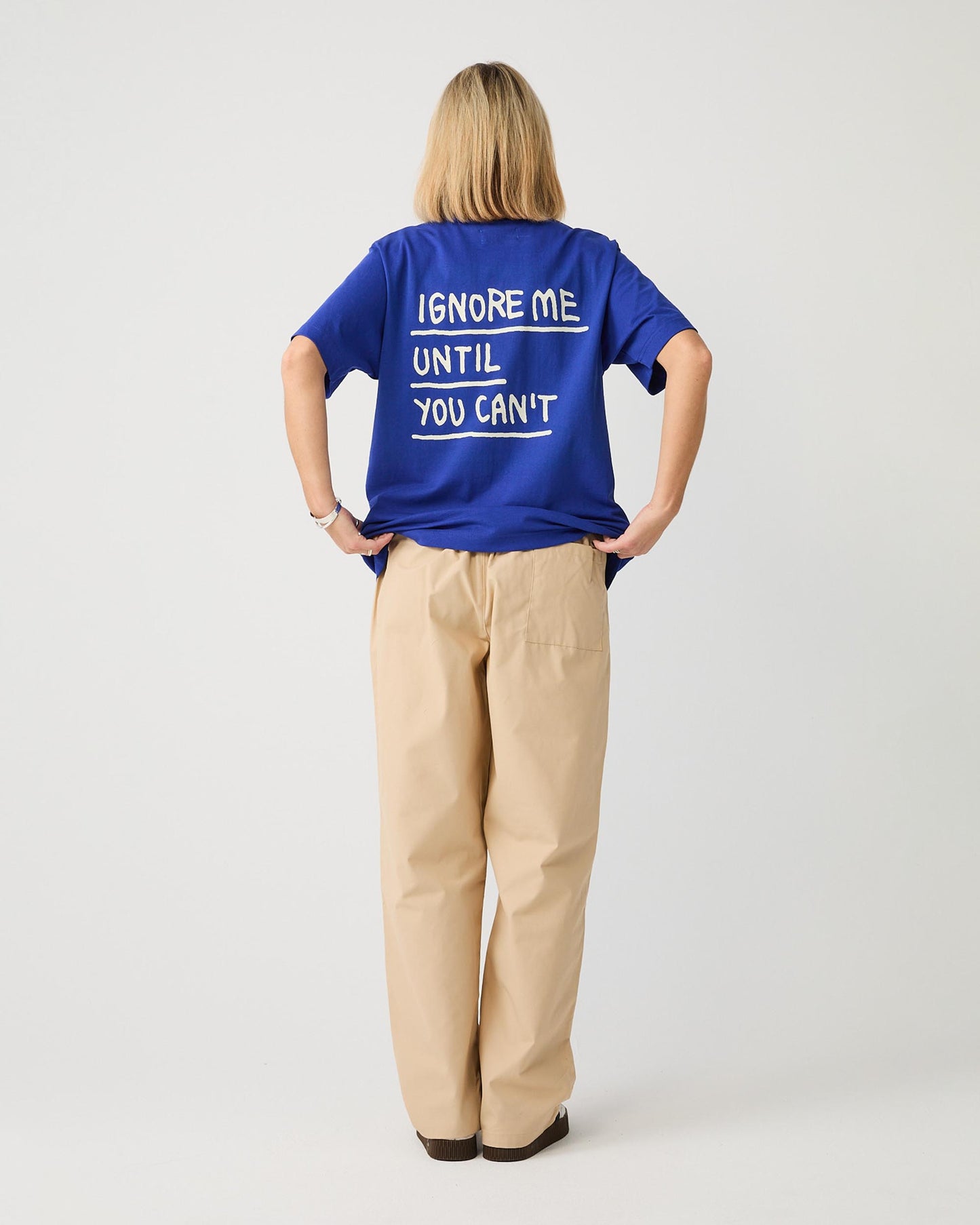 Kobaltblaues T-Shirt mit weißem Rückendruck, getragen von einer Person mit dem Rücken zur Kamera. Der großflächige Print zeigt den handschriftlich gestalteten Schriftzug „IGNORE ME UNTIL YOU CAN’T“, jeweils durch weiße Linien unterstrichen. Das Shirt hat einen lockeren Schnitt mit kurzen Ärmeln und wird über einer beigefarbenen Loungehose getragen.
