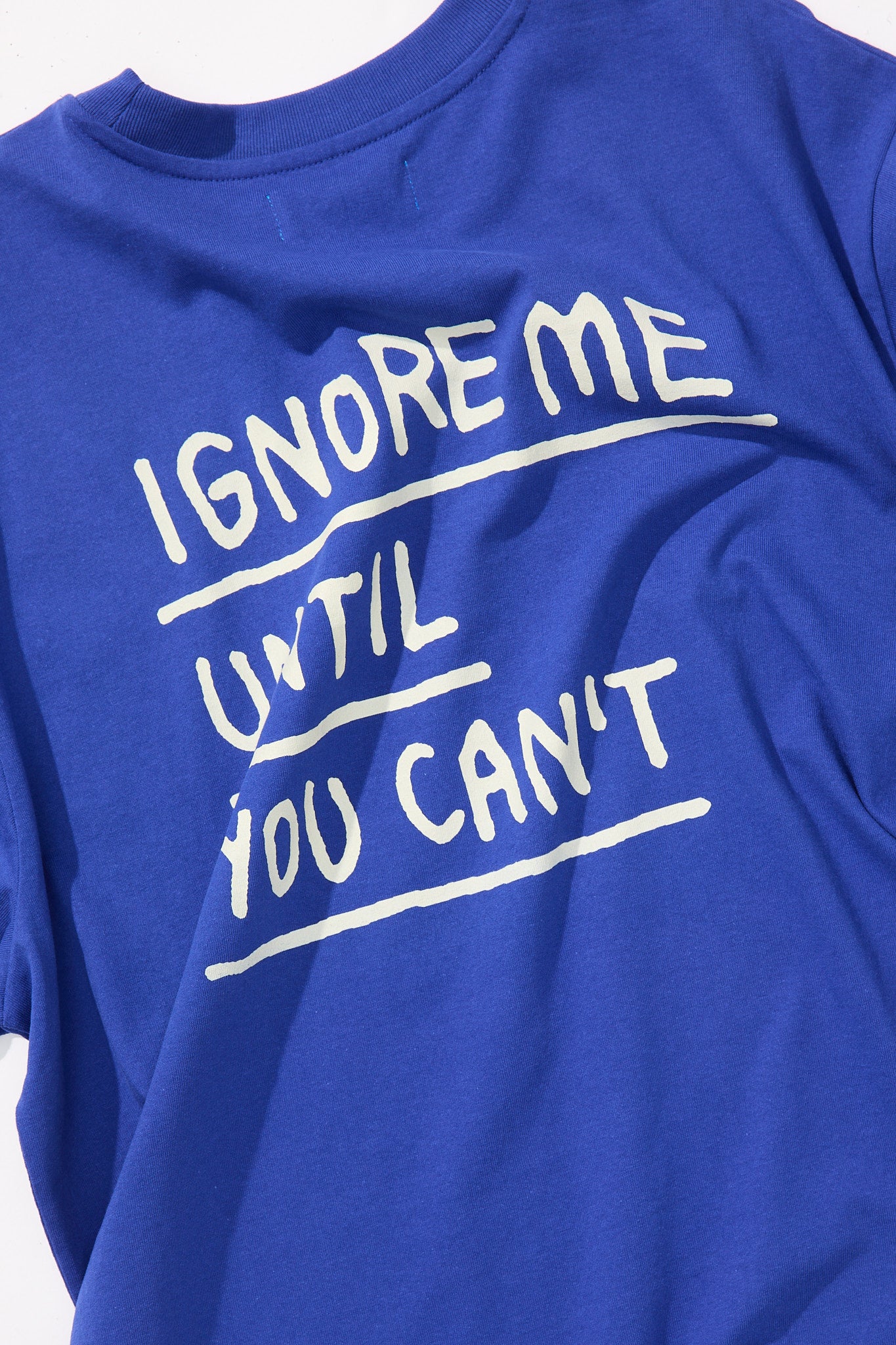 Nahaufnahme eines kobaltblauen T-Shirts mit weißem Rückendruck in handschriftlicher Optik. Der Print zeigt den markanten Schriftzug „IGNORE ME UNTIL YOU CAN’T“, wobei jede Zeile durch eine weiße Linie betont wird. Der Stoff ist leicht gewellt und zeigt feine Details der Textur.