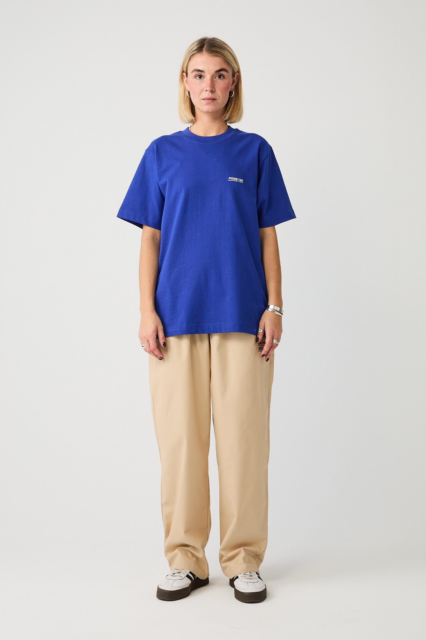Kobaltblaues T-Shirt mit lockerem Schnitt und weißem „PUSHER TONY“-Schriftzug auf der linken Brust, getragen von einer frontal stehenden Person. Das Shirt hat einen klassischen Rundhalsausschnitt und wird kombiniert mit einer sandfarbenen Loungehose und weißen Sneakers mit dunkler Sohle. Das Design wirkt clean und modern mit dezentem Markenfokus.