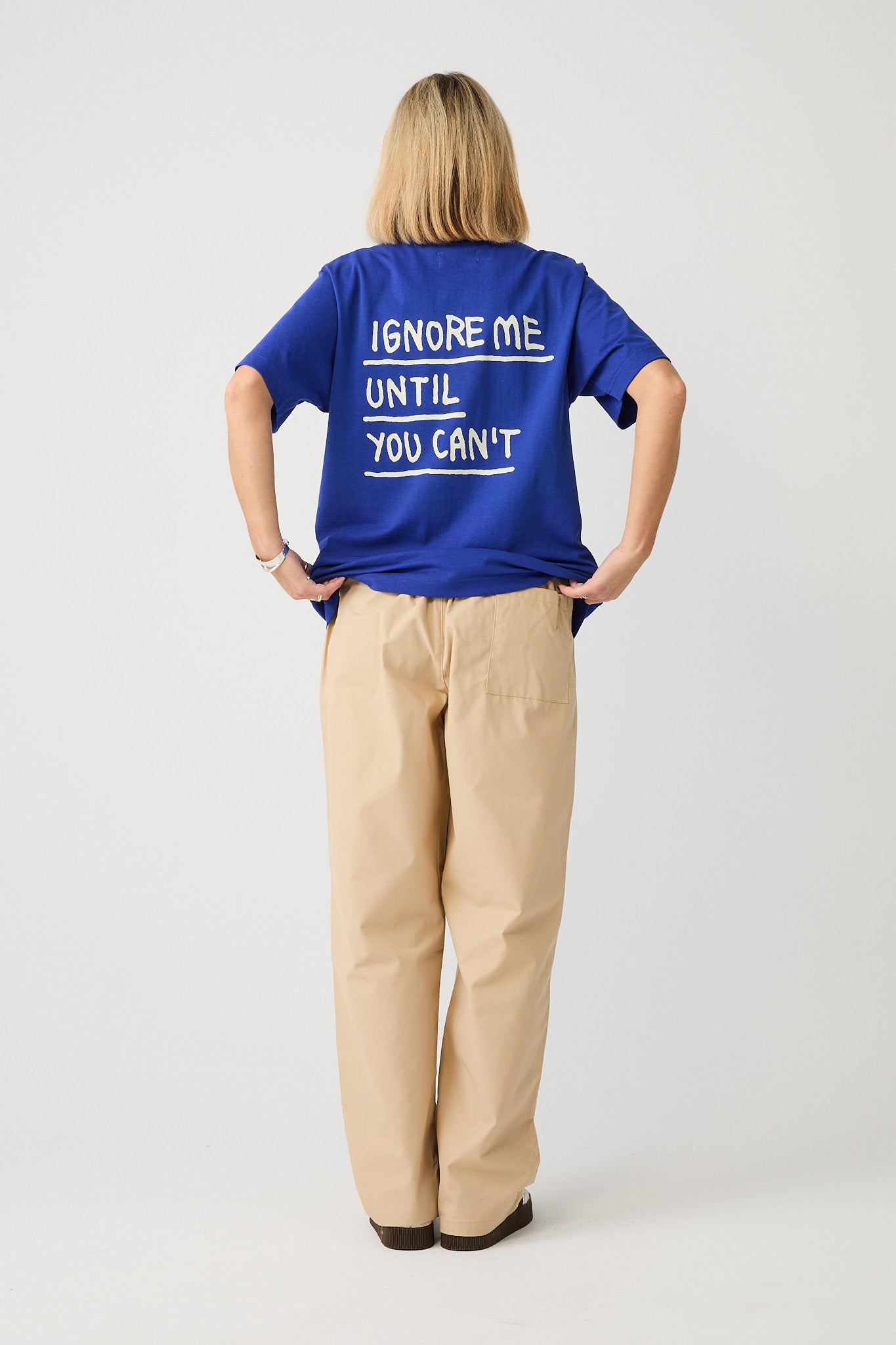 Kobaltblaues T-Shirt mit weißem Rückendruck, getragen von einer Person mit dem Rücken zur Kamera. Der großflächige Print zeigt den handschriftlich gestalteten Schriftzug „IGNORE ME UNTIL YOU CAN’T“, jeweils durch weiße Linien unterstrichen. Das Shirt hat einen lockeren Schnitt mit kurzen Ärmeln und wird über einer beigefarbenen Loungehose getragen.